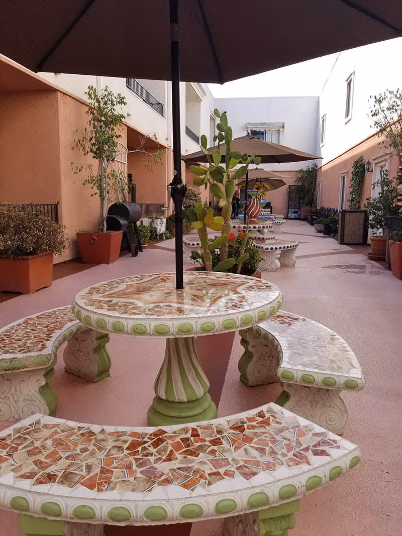 Outdoor courtyard area with mosaic-tiled round tables and curved benches, each with a large umbrella. The space is surrounded by peach-colored building walls with potted plants and small trees along the sides.