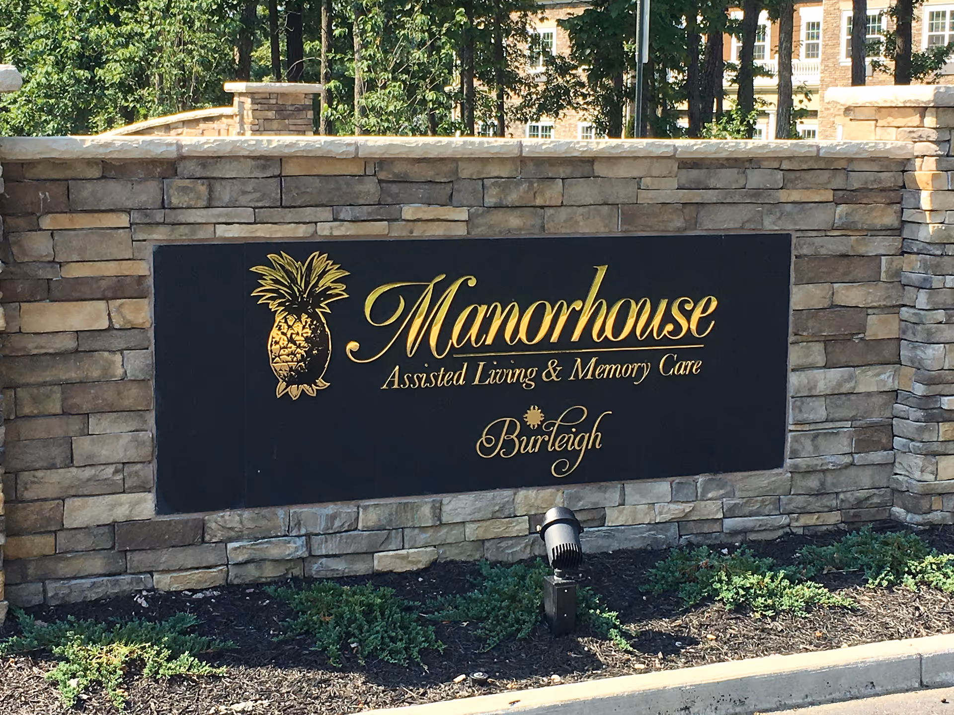 Stone wall with a black plaque featuring gold lettering and a pineapple logo. The plaque reads 'Manorhouse Assisted Living & Memory Care Burleigh'. There are some small green plants and a spotlight in front of the wall, with trees and a building visible in the background.
