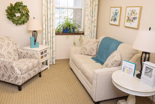 A cozy living room with a beige patterned armchair and a beige sofa adorned with patterned pillows and a teal throw blanket. There are two side tables with lamps, a small clock, and a decorative sign that says 'Be Kind'. A window with patterned curtains and a green potted plant is in the background. Two framed floral artworks hang on the wall.