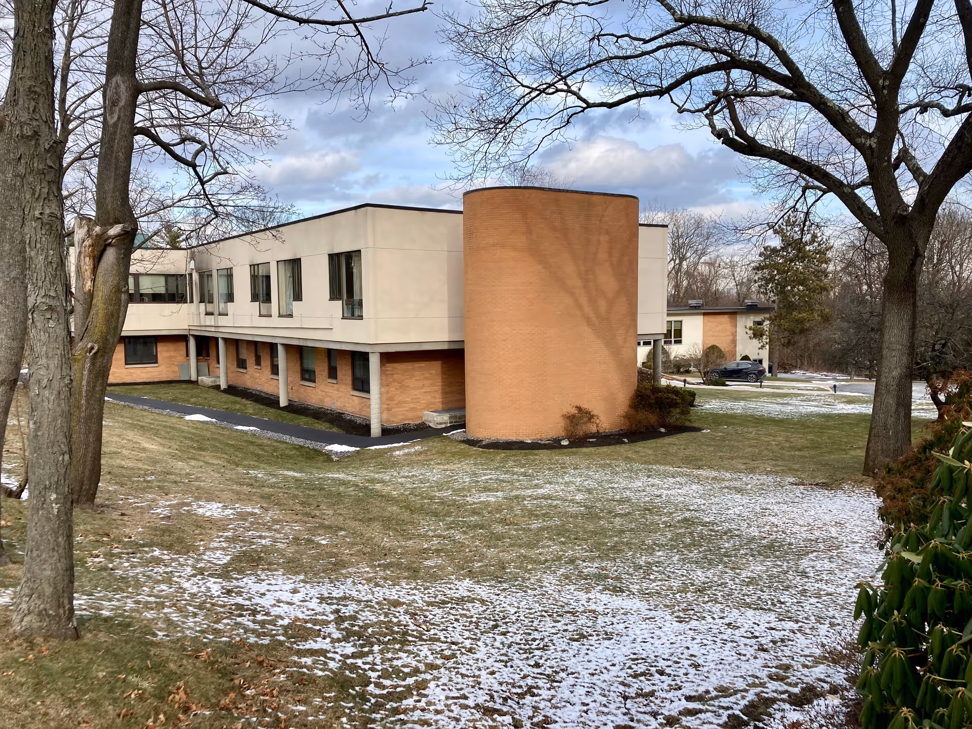 Exterior view of a two-story building with a mix of beige and brick walls, surrounded by a grassy area with patches of snow and several leafless trees under a partly cloudy sky.