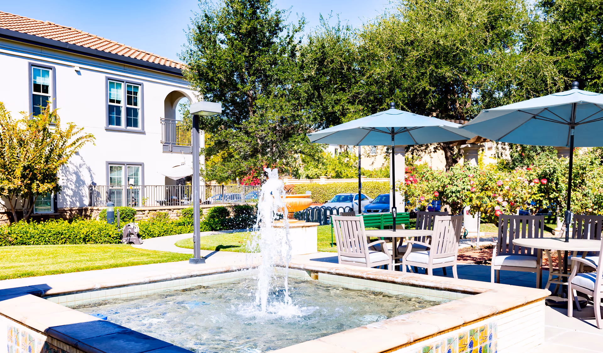 Outdoor courtyard area at Cogir of Brentwood featuring a water fountain in the center, surrounded by patio tables with umbrellas and chairs. There are trees, bushes, and a building with windows in the background under a clear blue sky.