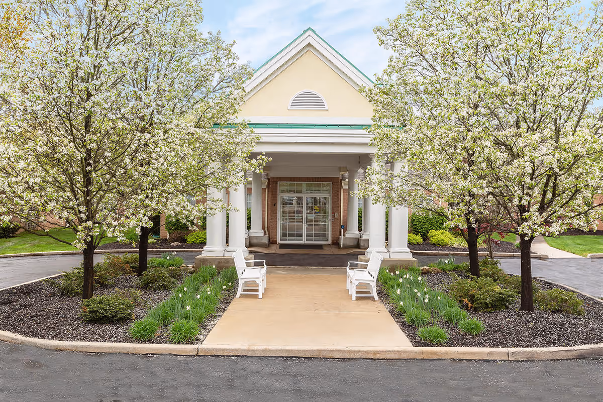 Entrance of Blossom Care Center of Cuyahoga Falls with a covered porch supported by white columns, flanked by two white benches and landscaped with blooming trees and green plants.