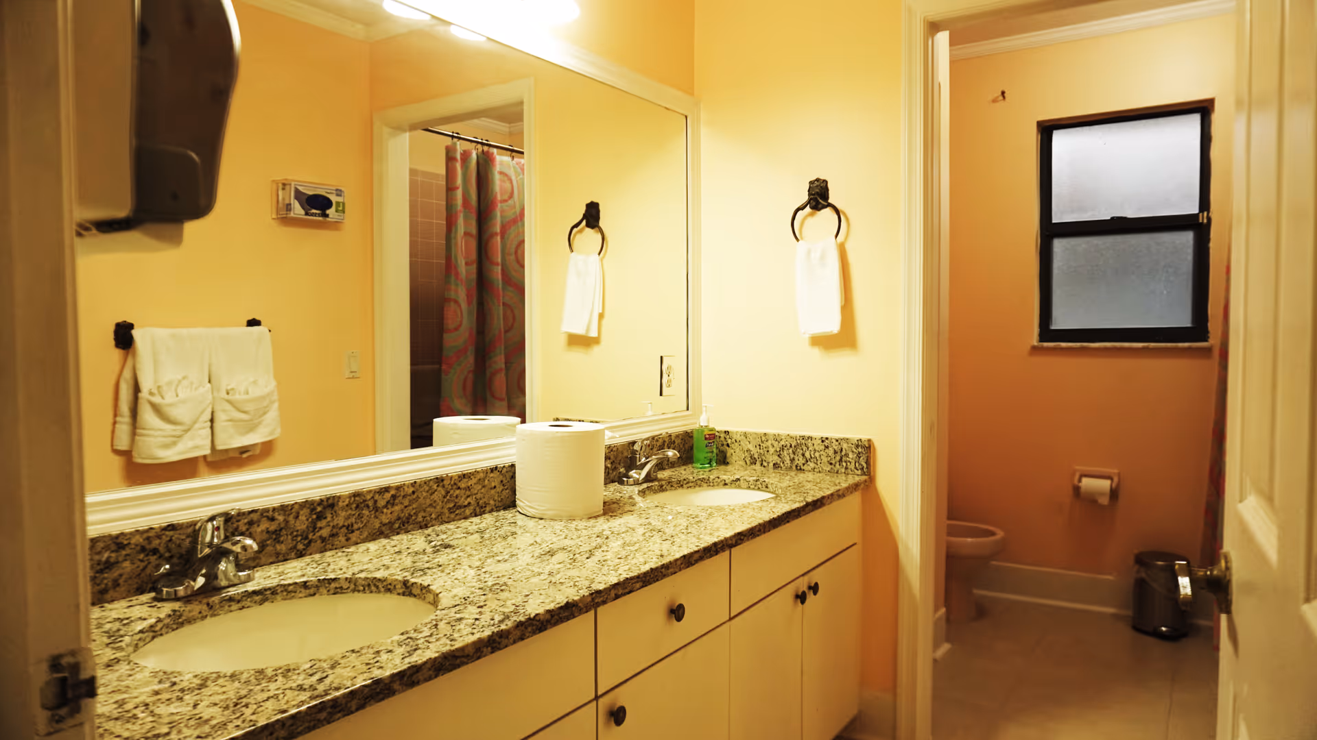 A bathroom with a double sink vanity featuring a granite countertop and two mirrors above. There are white towels hanging on black towel rings and a patterned shower curtain visible in the reflection. The bathroom has peach-colored walls and a frosted window above the toilet.