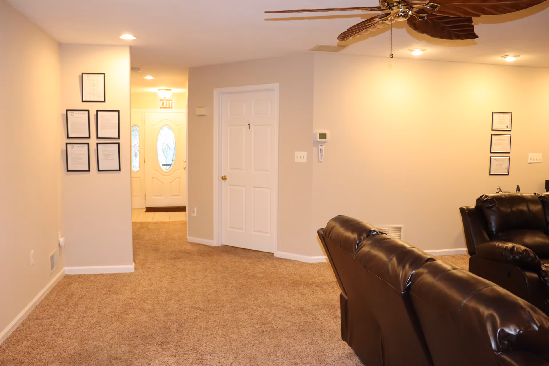 Interior view of a living area in an assisted living facility with beige walls and carpeted floor. There are two dark brown leather recliners on the right side, a white door labeled with the number 1 in the center, framed certificates on the walls, and a ceiling fan with leaf-shaped blades above.