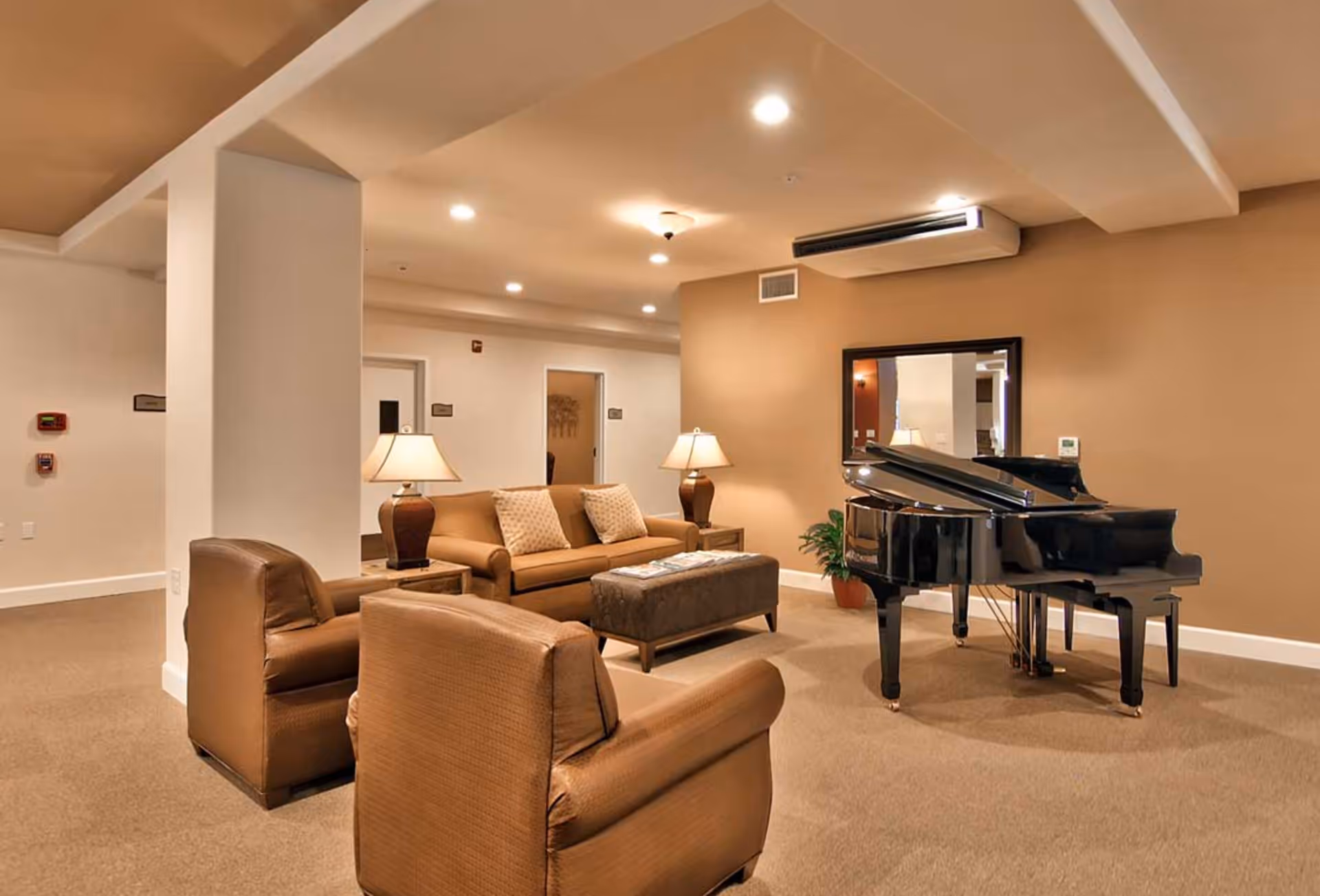A cozy senior living common area with beige walls and carpet, featuring a black grand piano, a brown sofa with two patterned cushions, two brown armchairs, two table lamps on side tables, a rectangular ottoman, a large mirror on the wall, and a potted plant.