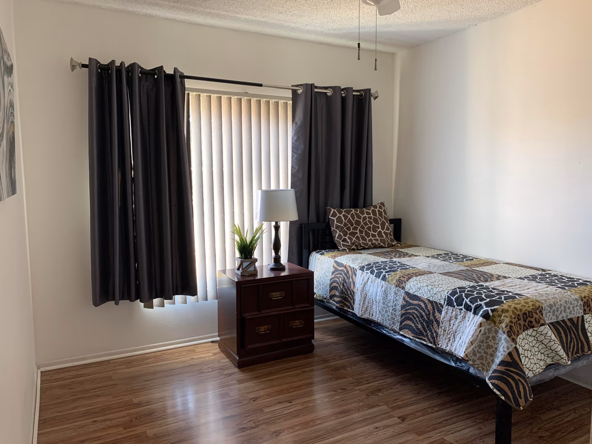Simple bedroom with a single bed covered by a patterned quilt, a nightstand with a lamp and plant, dark curtains over a window, and wood flooring.