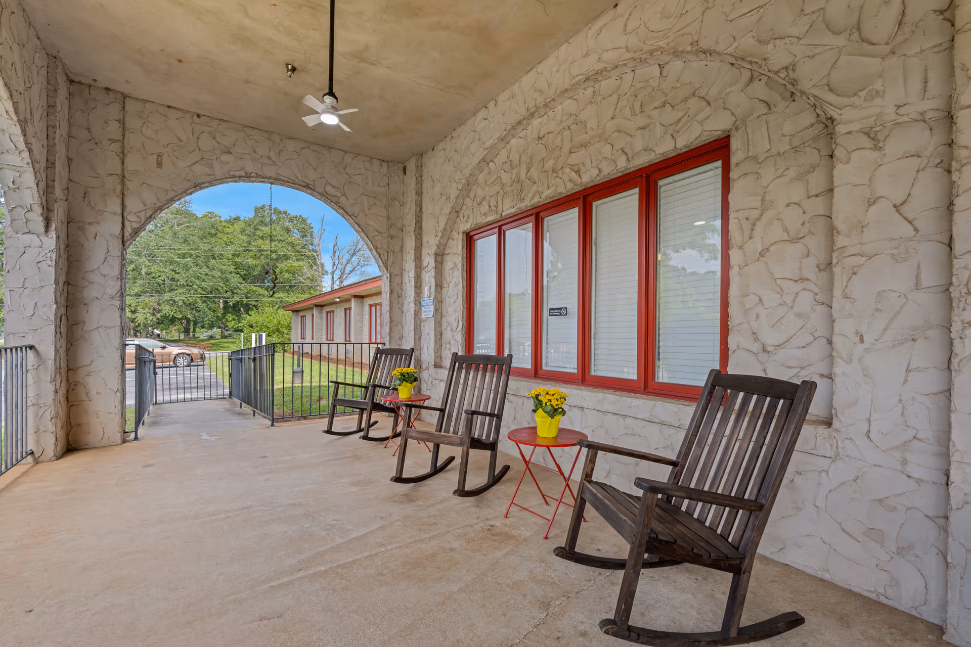 Covered front porch with three wooden rocking chairs and small red tables holding yellow potted flowers along a stucco building facade.