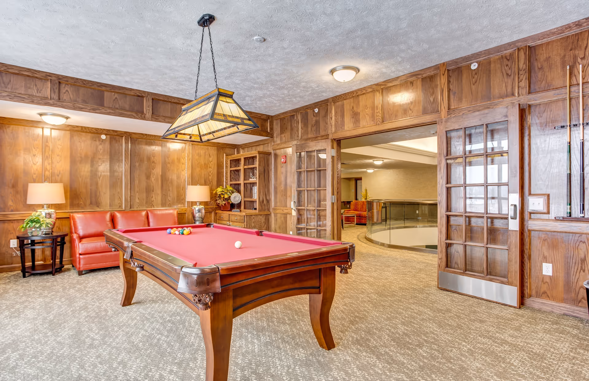 A cozy game room with a red felt pool table in the center, surrounded by wood-paneled walls. There is a red leather couch with two table lamps on either side, and a hanging stained glass light fixture above the pool table. The room has carpeted floors and a set of glass-paneled double doors leading to a hallway with additional seating and a railing overlooking a lower level.