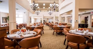 Spacious dining room with multiple round tables set with plates, glasses, and floral centerpieces. The room features high ceilings with chandeliers, large windows allowing natural light, and a warm, inviting atmosphere with beige walls and carpeted floors.