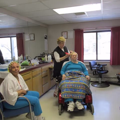 A hairdresser styling the hair of an elderly woman seated in a wheelchair covered with a colorful blanket, while another elderly woman with hair rollers sits nearby in a chair inside a well-lit room with large windows and red curtains.