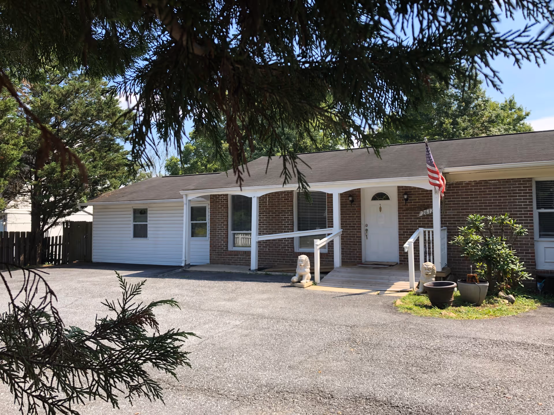 Front exterior view of a single-story brick and siding building with a small porch featuring a wheelchair ramp, two lion statues, potted plants, and an American flag. The building is surrounded by trees and has a paved driveway in front.