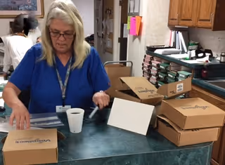 A woman in blue scrubs is working at a counter in a facility, handling paperwork and surrounded by several stacked cardboard boxes. The background shows office supplies and cabinets.