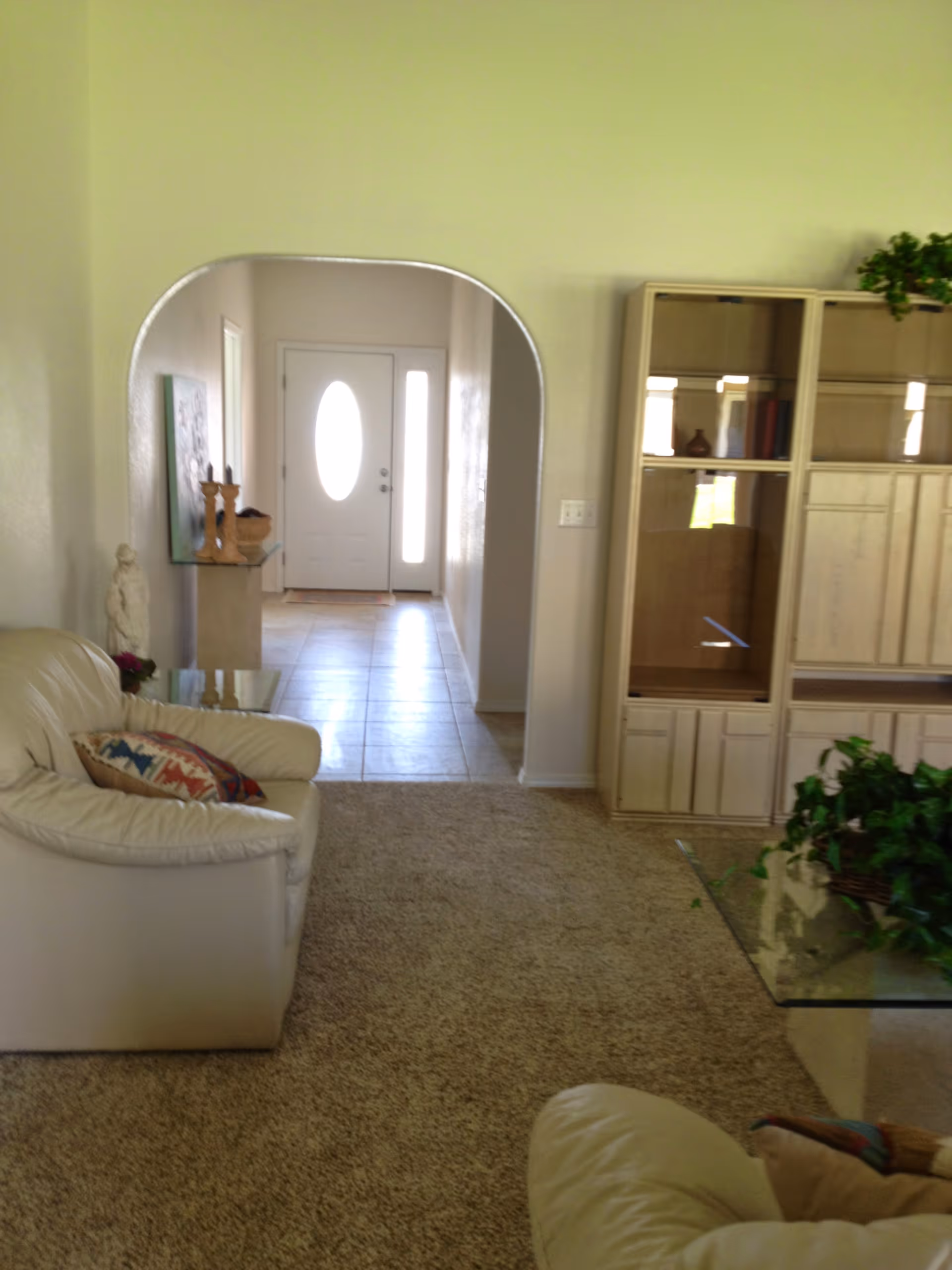 Living room with cream leather couches, a glass coffee table, built-in shelving and an arched hallway leading to a front door.