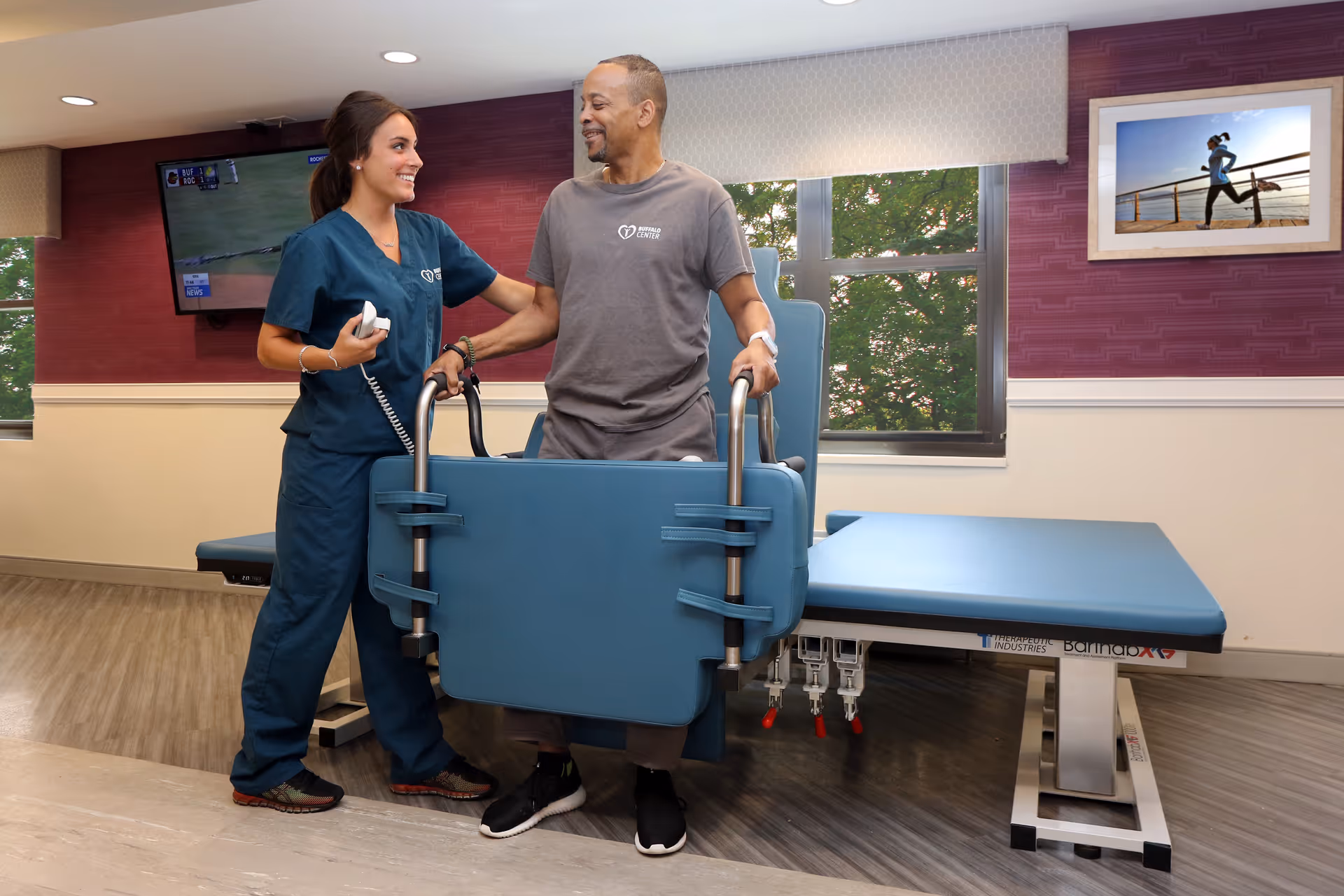 A healthcare worker in blue scrubs assists a man using a standing aid device in a rehabilitation room with a treatment table, windows showing greenery outside, a wall-mounted TV, and a framed picture of a person running on the wall.