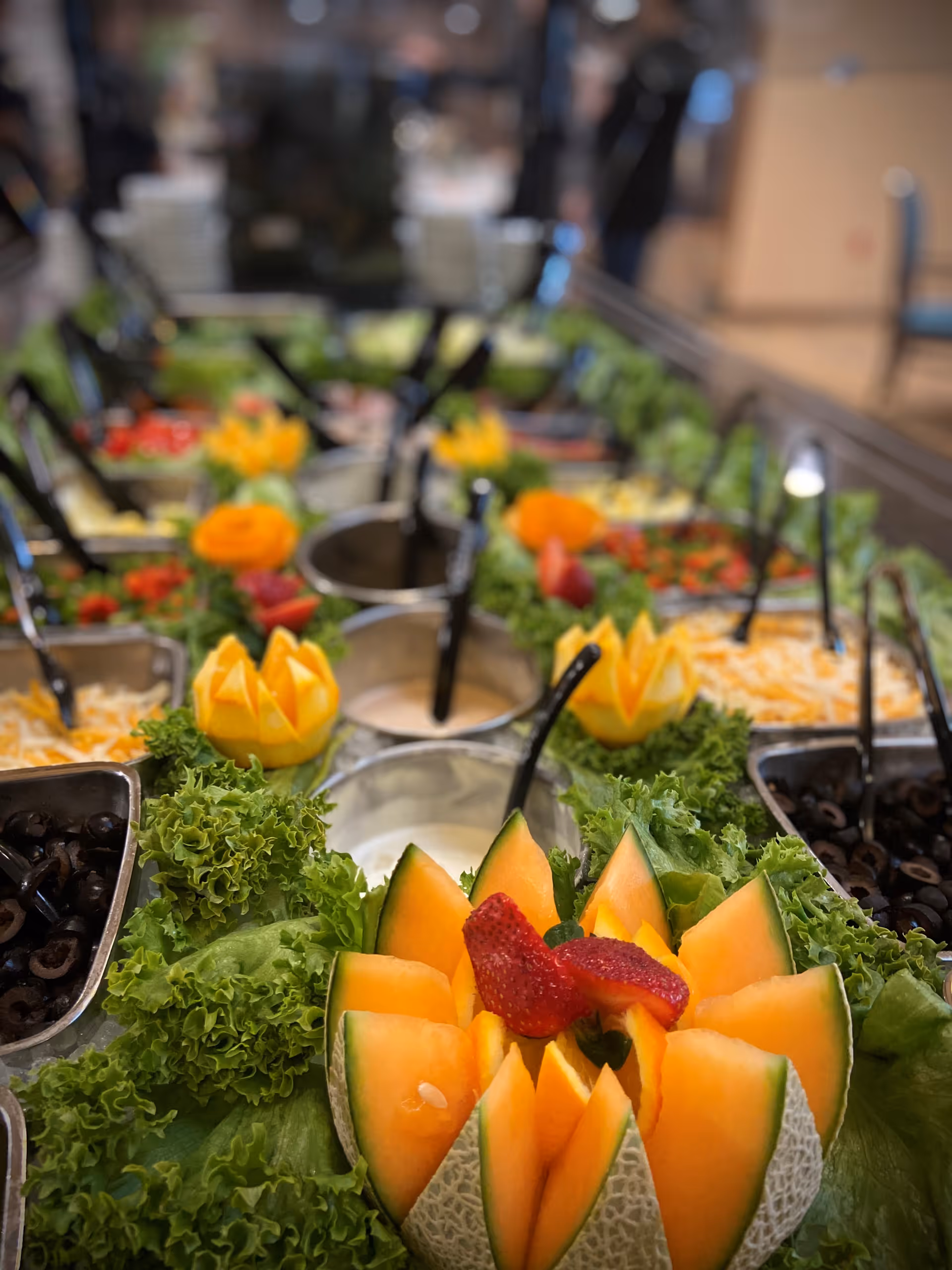 A close-up view of a salad bar featuring fresh green lettuce, sliced black olives, shredded cheese, various dressings, and decorative fruit carvings including cantaloupe slices with strawberries on top.