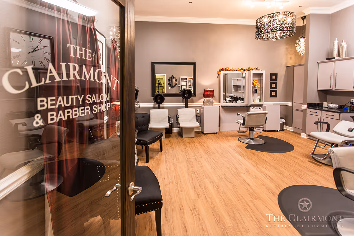 Interior view of The Clairmont Retirement Community's beauty salon and barber shop featuring salon chairs, hair washing stations, mirrors, and modern lighting fixtures with wooden flooring and neutral-colored walls.
