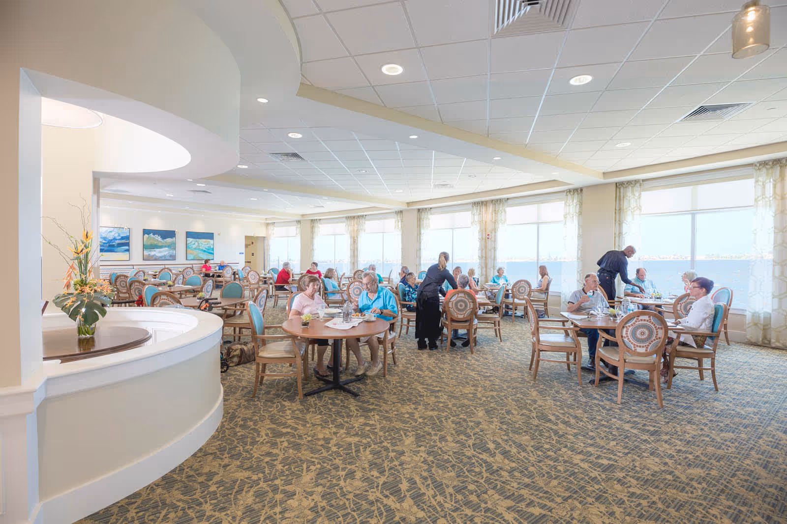 A bright and spacious dining room in a senior living facility with large windows overlooking a body of water. Several elderly residents are seated at round tables with wooden chairs, enjoying meals and socializing. A staff member is assisting one of the tables. The room has light-colored walls, patterned carpet, and decorative paintings on the far wall.