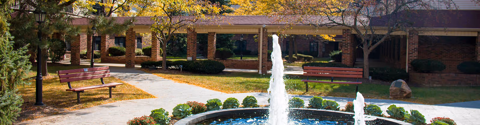 Outdoor courtyard area at a senior living facility with a circular fountain in the center, surrounded by plants and flowers. There are two wooden benches on either side of the paved walkway, with brick buildings and a covered walkway in the background. Trees with autumn foliage are visible around the area.