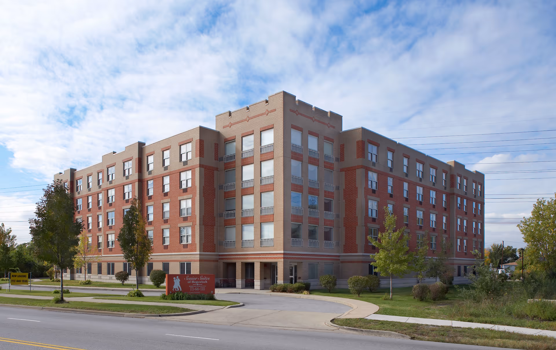 Exterior view of a four-story brick senior living facility named Senior Suites Of Hegewisch, with a driveway and some trees in front under a partly cloudy sky.
