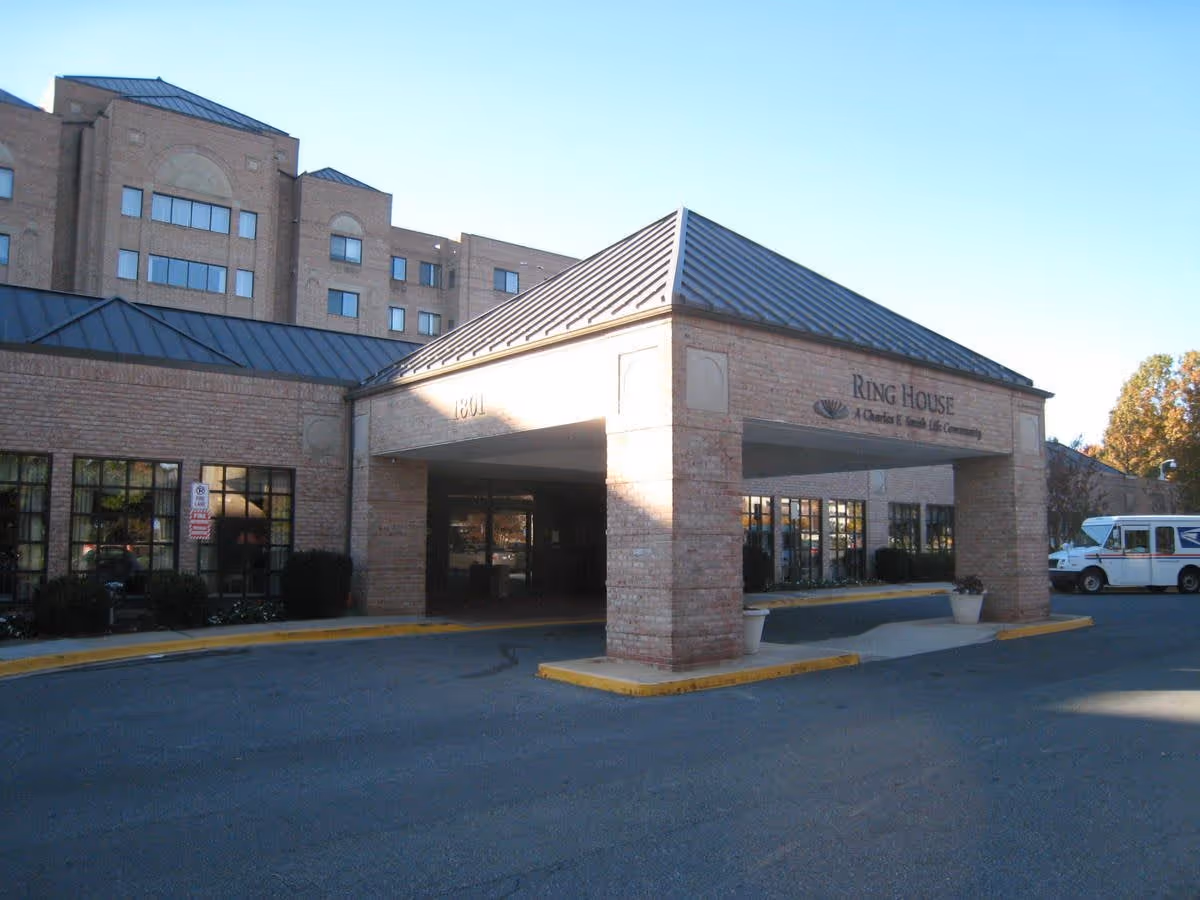 Exterior view of the entrance to Ring House, a senior living community, showing a covered drop-off area with brick pillars and a metal roof. The building is multi-story with numerous windows, and a white postal service truck is parked nearby.