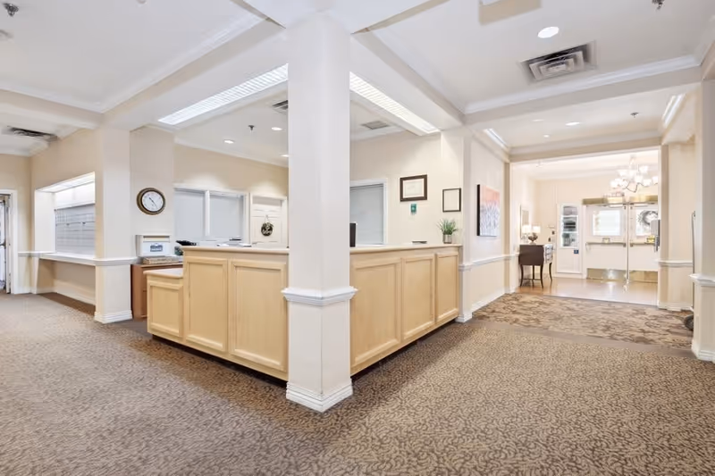 Interior view of a senior living facility reception area with a wooden front desk, beige walls, patterned carpet, a clock on the wall, framed certificates, a small plant on the desk, and double glass doors leading outside.