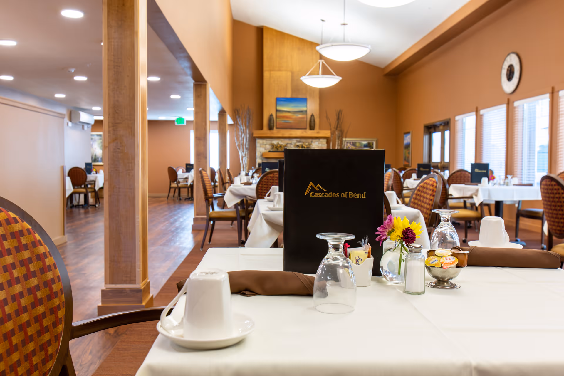 Warm wood-accented dining room with set tables, a menu labeled 'Cascades of Bend', flowers, and place settings.