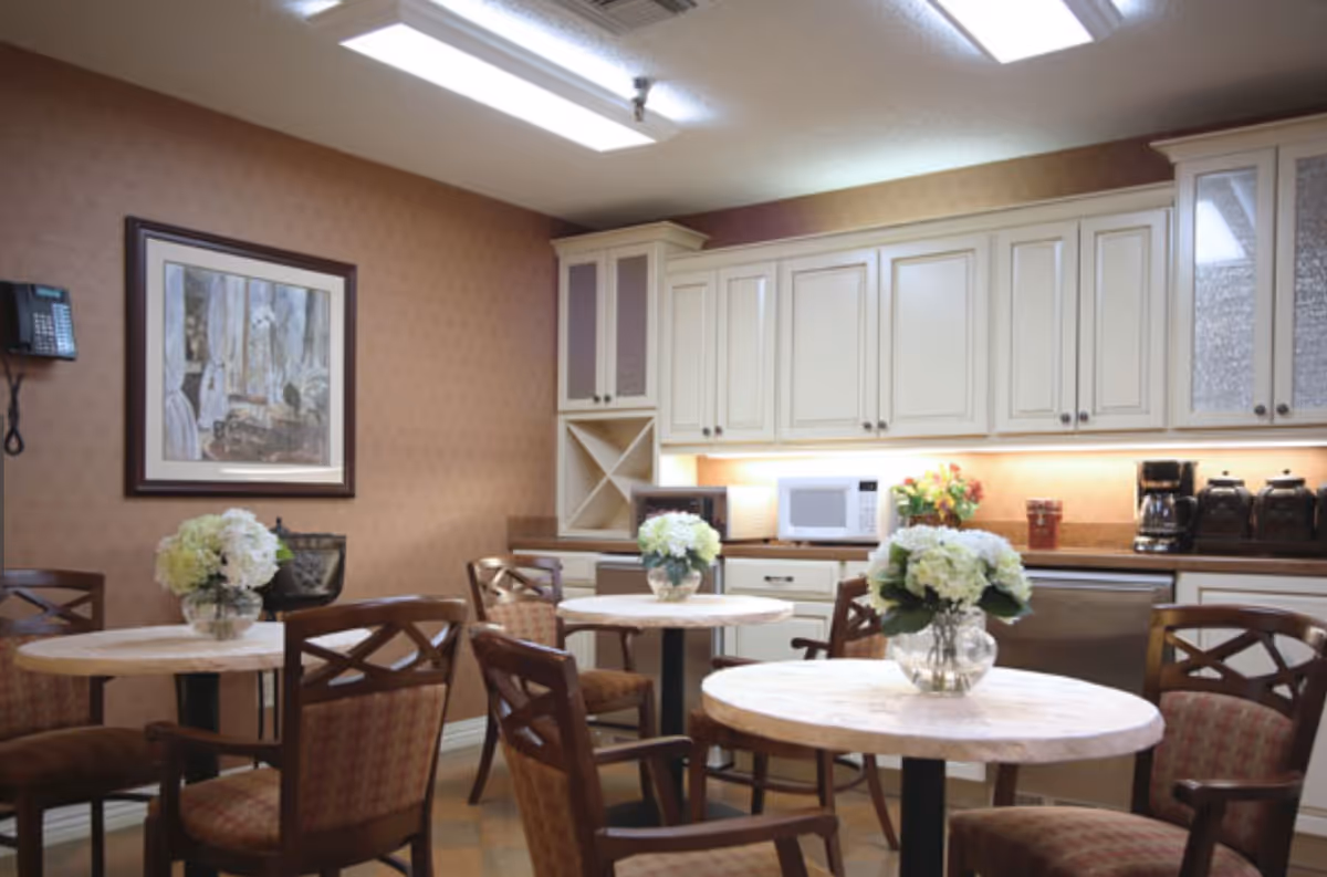 A cozy dining area in a senior living facility with round tables, each adorned with a vase of white flowers. The room features wooden chairs with cushioned seats, beige cabinetry with a microwave, coffee maker, and other kitchen appliances along the back wall. A framed painting and a wall-mounted telephone are visible on the left wall.