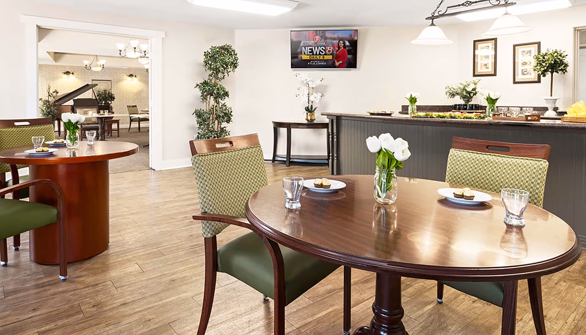 A bright and clean dining area with round wooden tables and green cushioned chairs. Each table has a small vase with white flowers and plates with cupcakes. In the background, there is a counter with food platters and decorative plants, a wall-mounted TV displaying a news channel, and an adjacent room with a grand piano and additional seating.