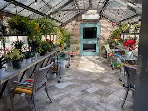 Interior view of a bright greenhouse room with a glass ceiling and brick walls. The space contains multiple tables and chairs with colorful cushions, surrounded by various potted plants and flowers. A teal-colored door with a window is visible at the far end of the room.