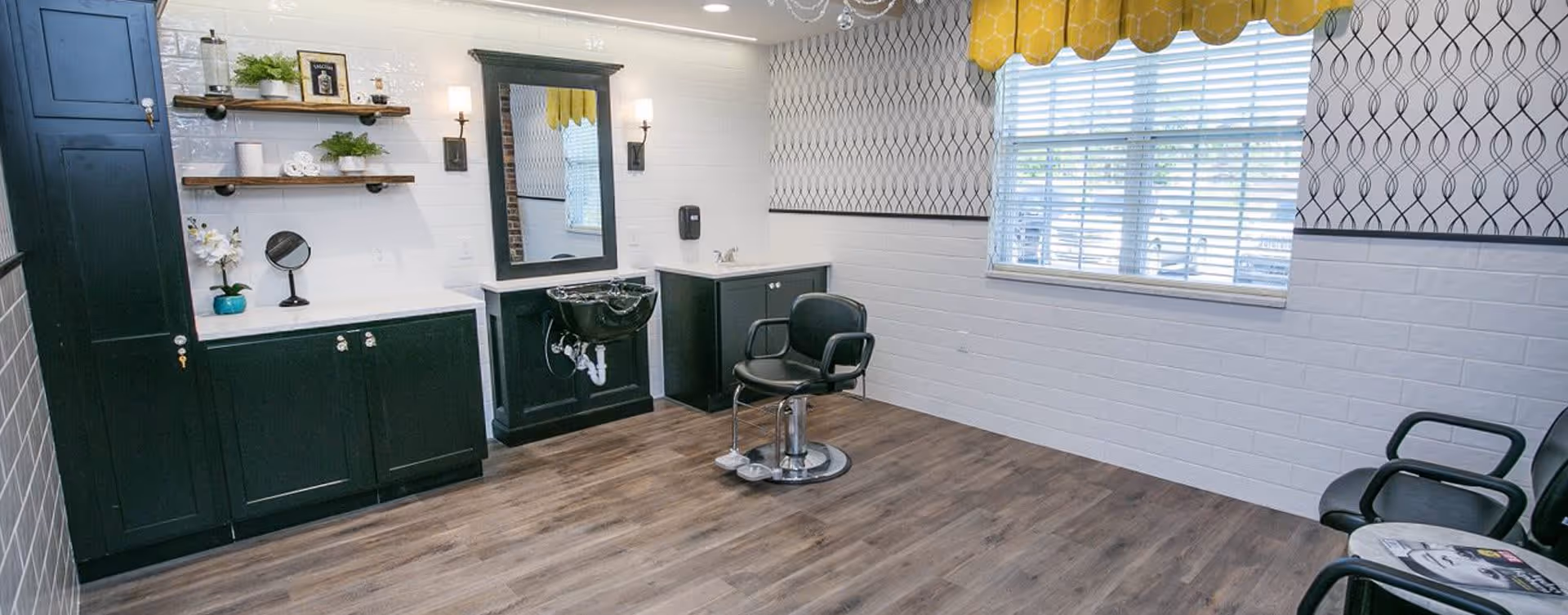 A clean and modern hair salon area with a black salon chair in front of a large mirror and a black sink. The room has dark wood flooring, white brick walls with a patterned wallpaper border near the ceiling, and a window with blinds and a yellow valance. There are black cabinets and shelves with decorative items on the left side, and additional black chairs on the right side.