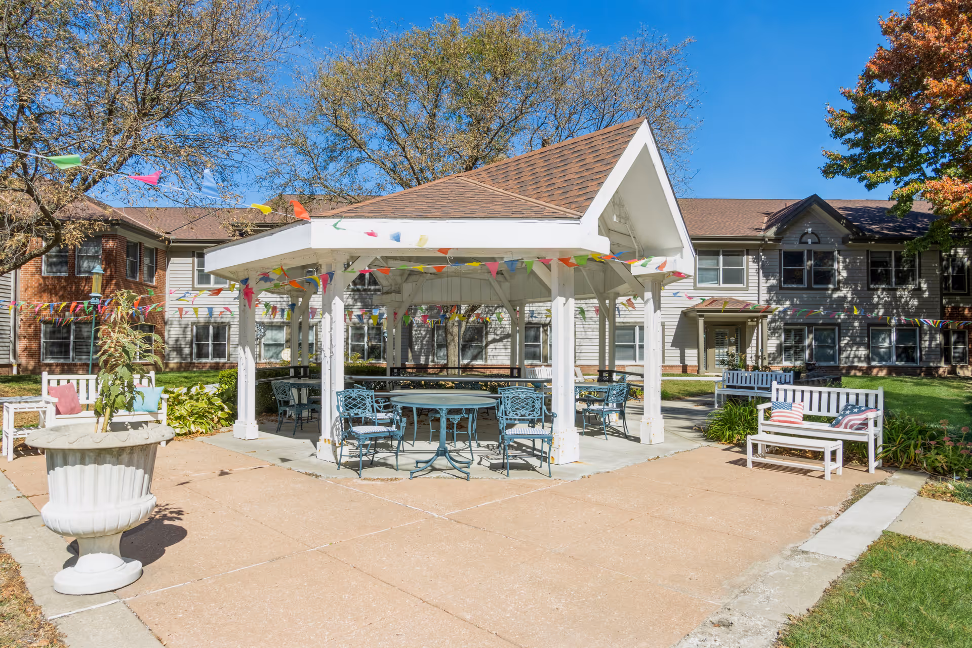 Outdoor patio area at The Forum at Overland Park featuring a white gazebo with a brown shingled roof, surrounded by metal tables and chairs. Colorful triangular pennant flags are strung around the gazebo. White benches with cushions and a large white planter with a plant are also visible. The background shows a two-story building with windows and trees with autumn foliage under a clear blue sky.