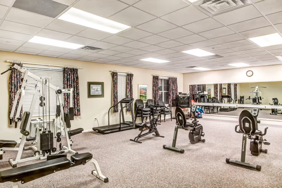 A senior living facility exercise room with various fitness equipment including a multi-station weight machine, two recumbent exercise bikes, and a treadmill. The room has beige walls, floral curtains on the windows, framed artwork, a large wall mirror, and a carpeted floor. Several stacked chairs and a small TV are also visible.