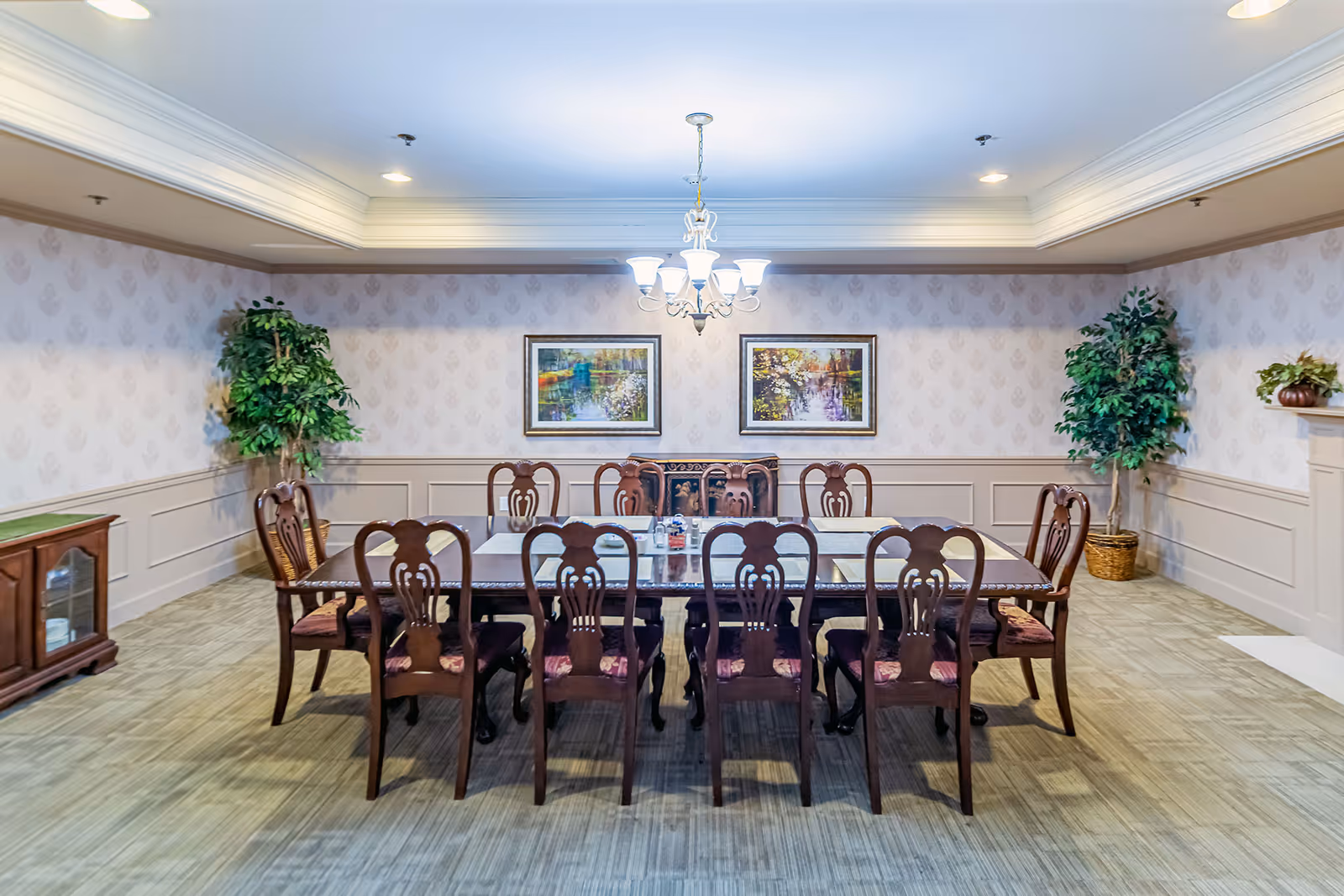 A formal dining room with a long rectangular wooden table surrounded by ten matching wooden chairs with cushioned seats. The room has patterned wallpaper, two framed landscape paintings on the wall, two large potted plants in the corners, a chandelier hanging from the ceiling, and a wooden cabinet on the left side.