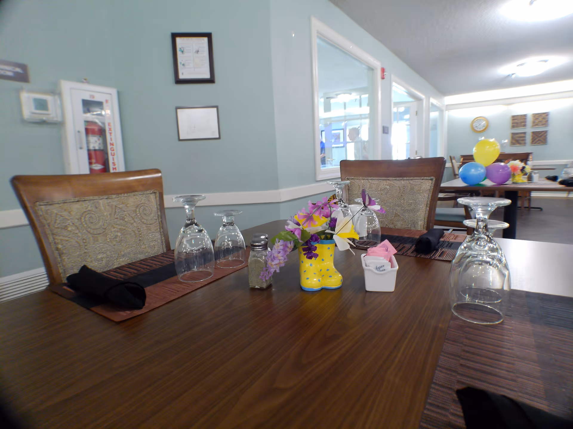 A dining table set with upside-down wine glasses, black cloth napkins, a small salt shaker, and a decorative yellow boot-shaped vase with purple and yellow flowers. In the background, there are more tables with chairs, colorful balloons, and a clock on the wall in a well-lit room with light blue walls and framed pictures.