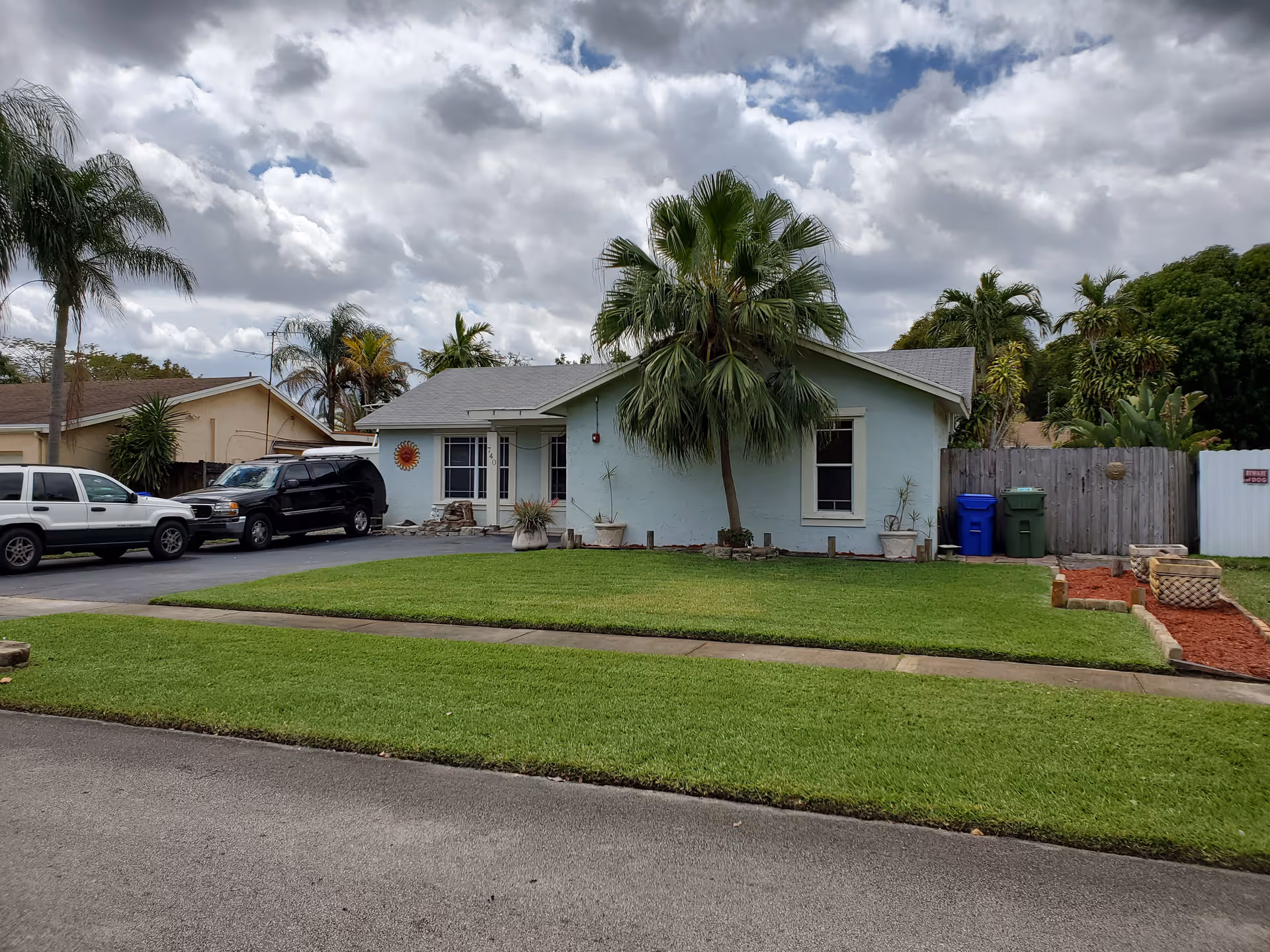 Single-story light blue house with a gray roof, a palm tree in front, and a well-maintained green lawn. There are two vehicles parked in the driveway to the left of the house. The sky is cloudy with patches of blue. There are various plants and a wooden fence on the right side of the house.