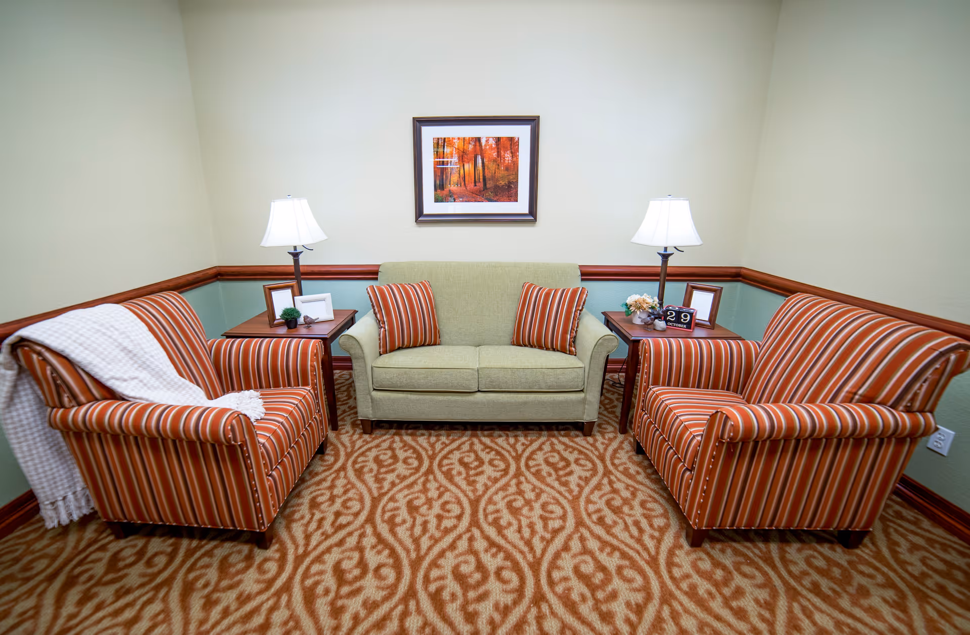 A cozy sitting area with a green loveseat flanked by two striped armchairs in shades of orange and brown. Each armchair has a side table with a lamp, picture frames, and small decorative items. A framed picture of an autumn forest scene hangs on the wall above the loveseat. The room has a patterned carpet in warm tones and light green walls with wooden trim.