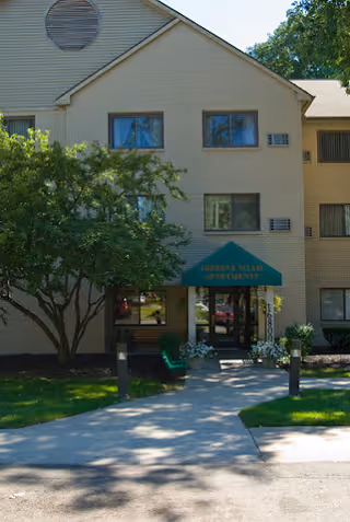 Front entrance of a beige brick apartment building with a green awning reading "Theresa Maxis Apartments", a tree to the left, and a walkway leading to the door.