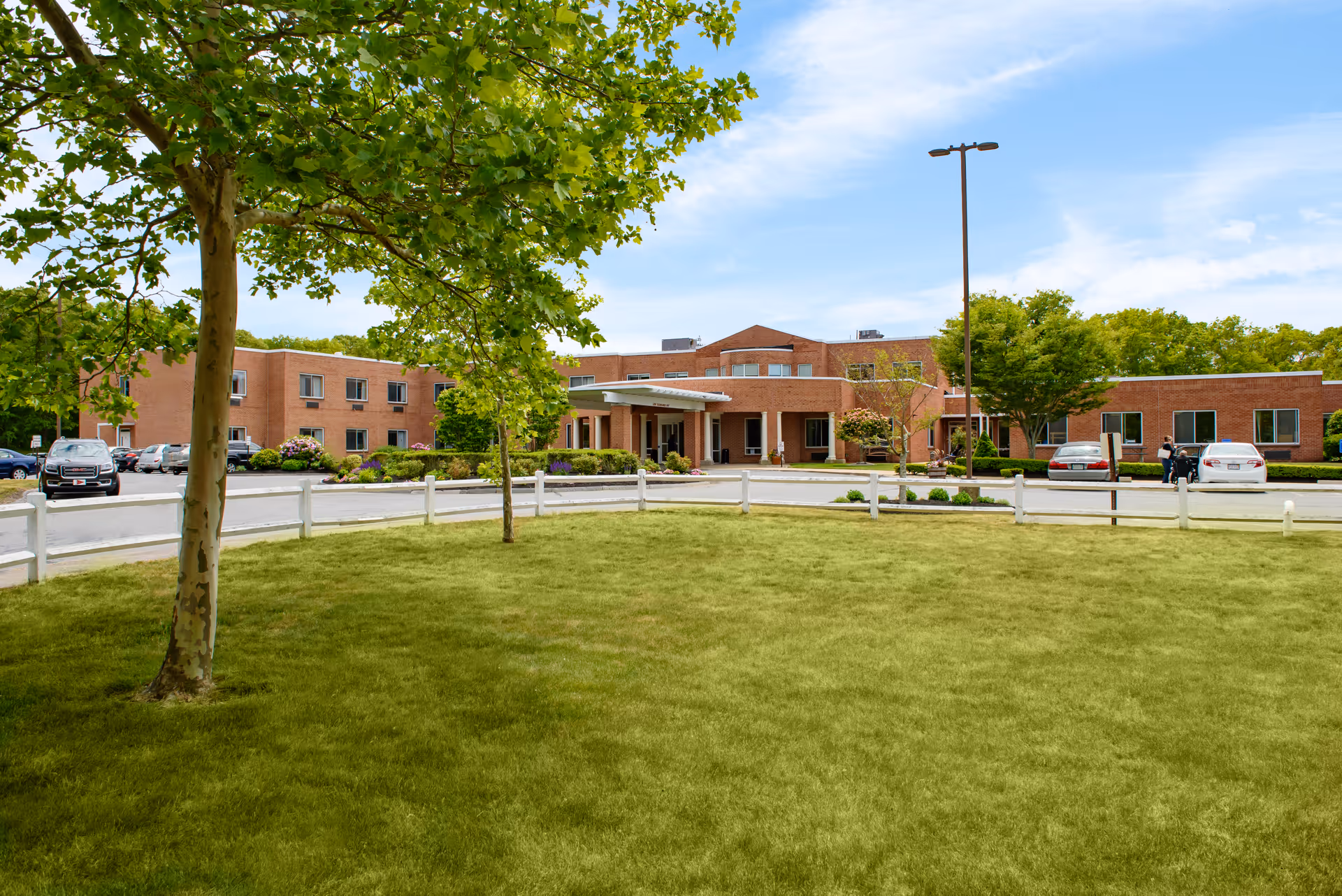 Front exterior view of a brick senior living facility with a covered entrance, surrounded by green grass, trees, and a white fence. Several cars are parked near the building under a partly cloudy sky.