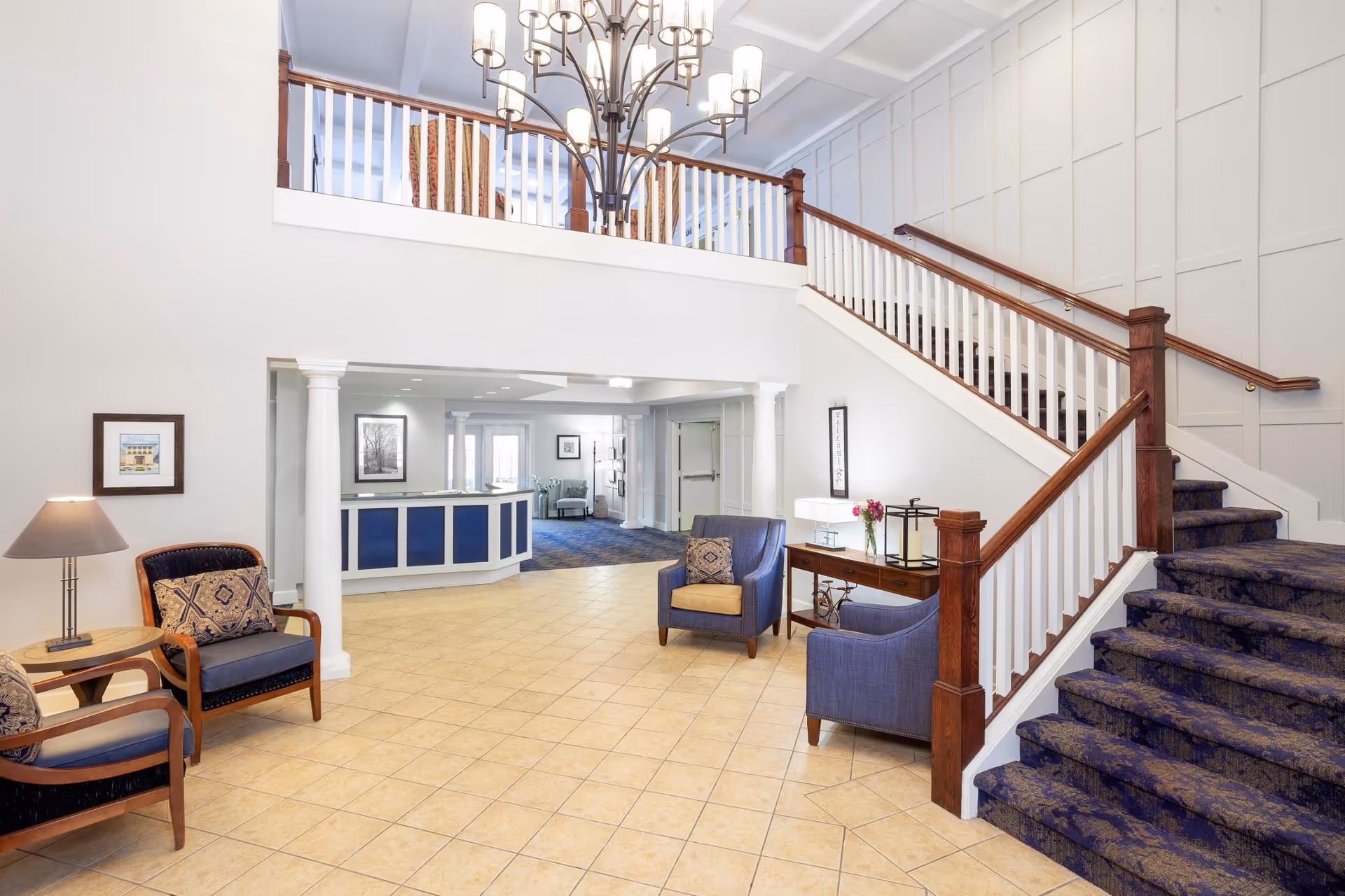 Interior view of a senior living facility lobby with a staircase on the right, blue and beige chairs with cushions, a wooden side table with a lamp and flowers, a reception desk in the background, and a large chandelier hanging from the ceiling.