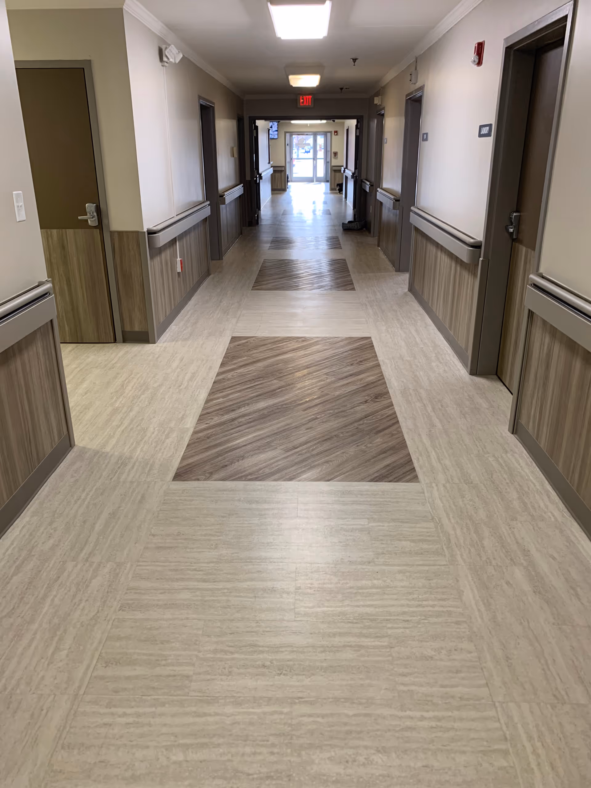 A clean, well-lit hallway in a senior living facility with beige walls, wood paneling halfway up the walls, and a patterned floor with alternating light and dark wood-like tiles. Several closed doors line both sides of the hallway, and an exit sign is visible at the far end near a glass door.