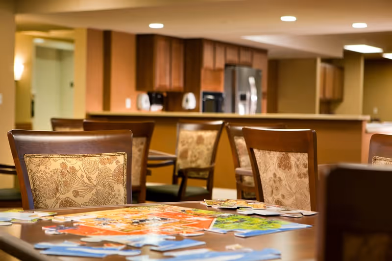 A cozy common area with a table in the foreground covered with puzzle pieces. Several wooden chairs with patterned cushions surround the table. In the background, there is a kitchen area with wooden cabinets, a refrigerator, and a counter. The lighting is warm and inviting.