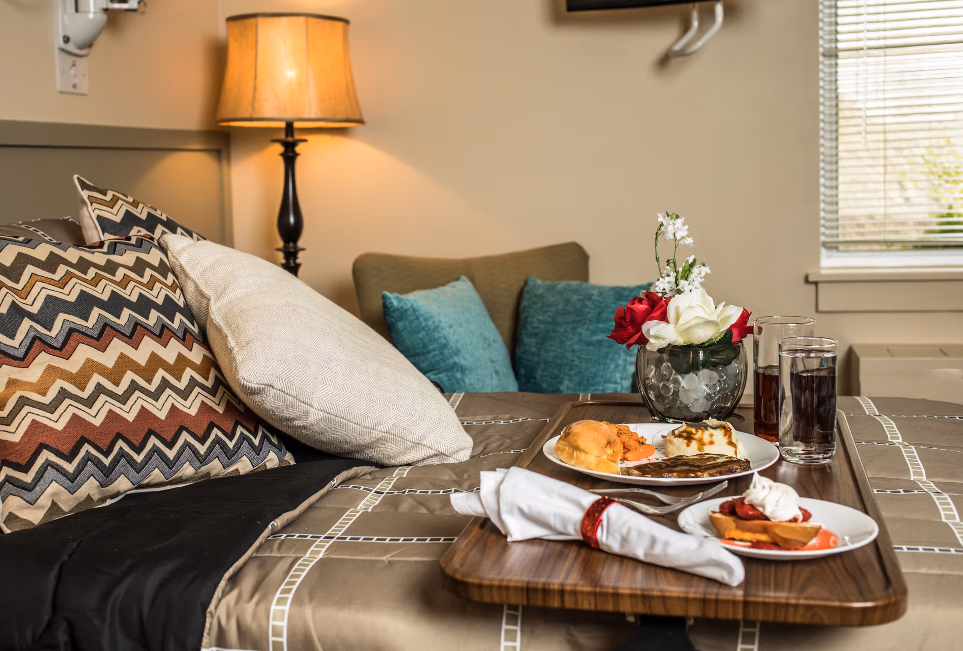 A cozy room with a bed featuring patterned and plain pillows, a bedside lamp, and a tray on the bed holding a meal with meat, mashed potatoes, carrots, a dessert, two glasses of beverages, and a small vase with red and white flowers.