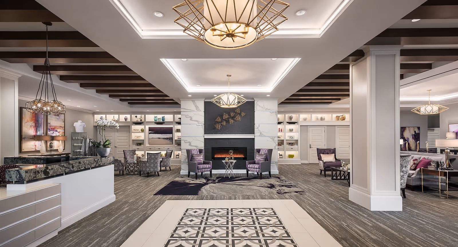 A spacious and elegantly designed senior living facility common area with a modern fireplace in the center, flanked by two purple armchairs. The room features patterned carpet, decorative ceiling lights, built-in shelves with art pieces and books, and a reception desk on the left side. The color scheme includes neutral tones with purple accents.