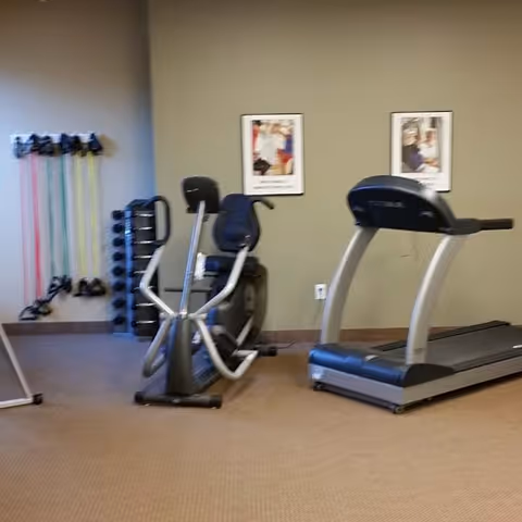 Exercise room with a treadmill, a recumbent exercise bike, resistance bands hanging on the wall, and a rack of dumbbells. Two framed pictures are hung on the beige wall behind the equipment.
