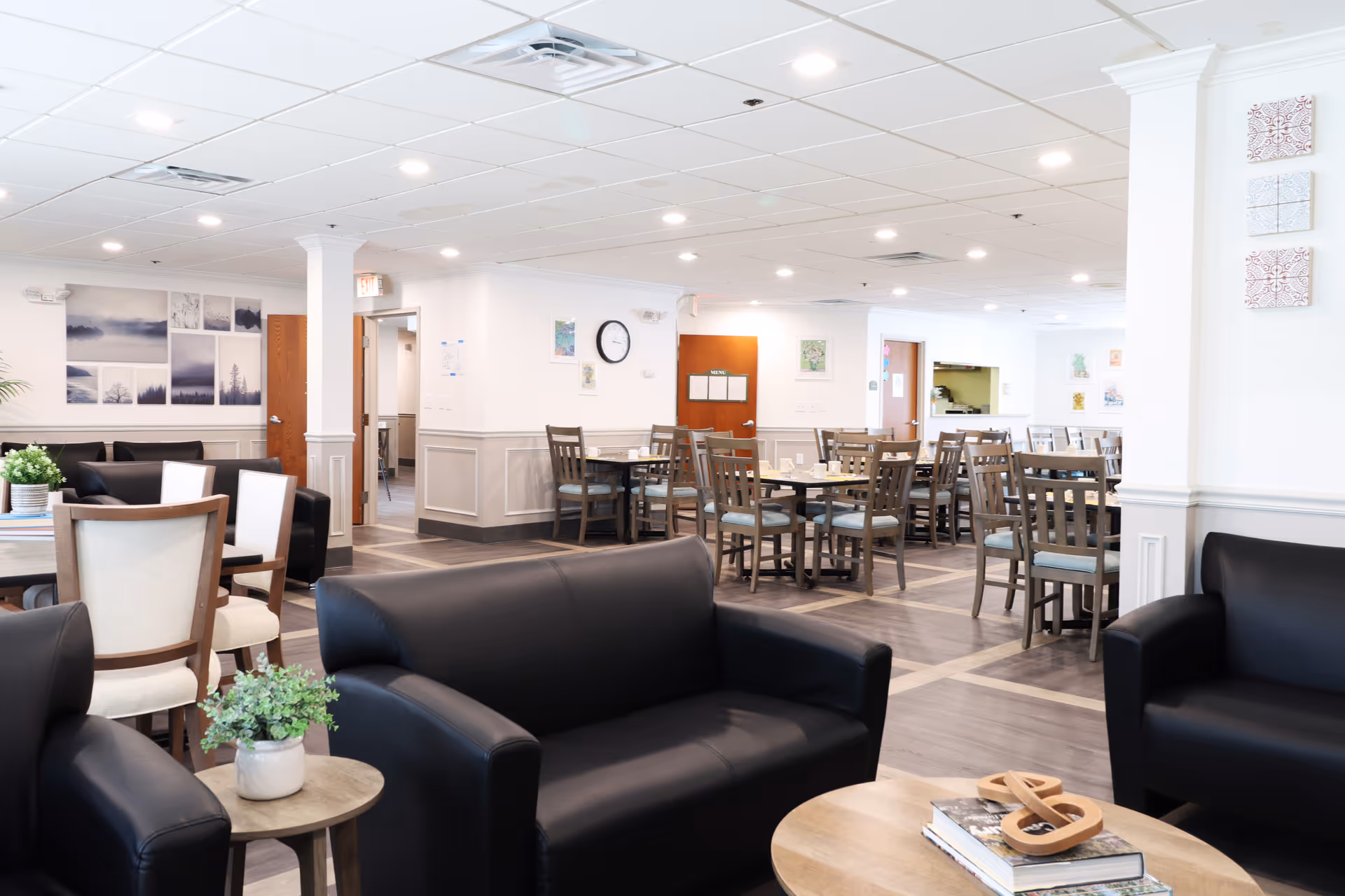 A bright and spacious common area in a senior living facility featuring black leather sofas and wooden chairs around tables. The room has white walls with decorative molding, framed artwork, and a clock. The flooring is a combination of wood and tile patterns, and the ceiling has recessed lighting.