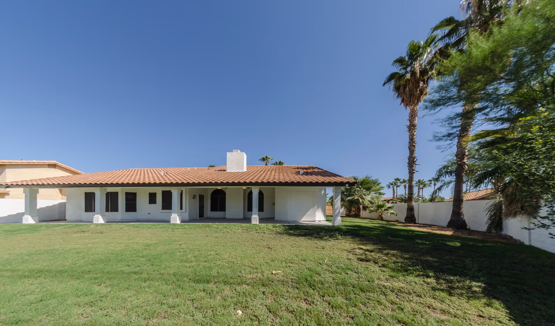 Single-story house with a red tile roof and white exterior walls, surrounded by a green lawn and palm trees under a clear blue sky.