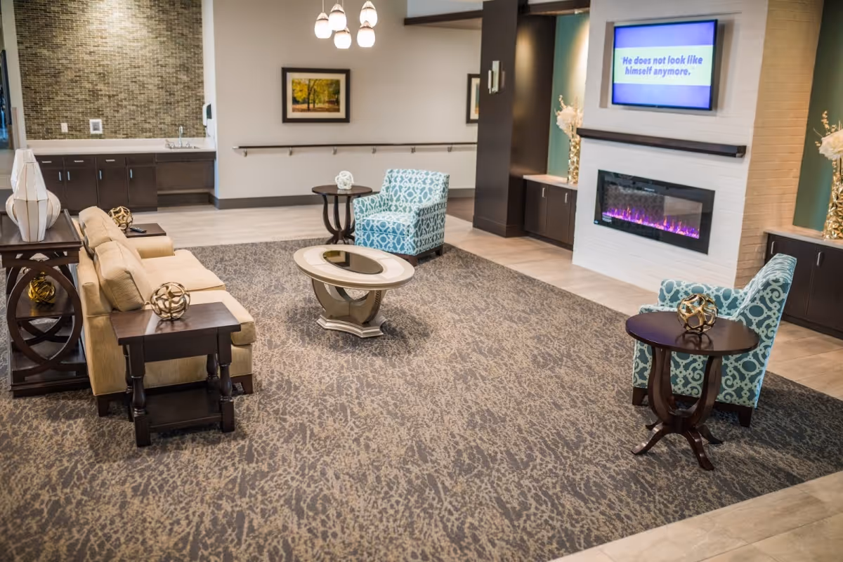 A cozy living room area with a beige sofa, two patterned blue armchairs, a round coffee table, and side tables with decorative items. The room features a modern electric fireplace with purple flames beneath a wall-mounted TV displaying the text 'He does not look like himself anymore.' The walls have framed artwork and the floor is covered with a patterned carpet and tile.
