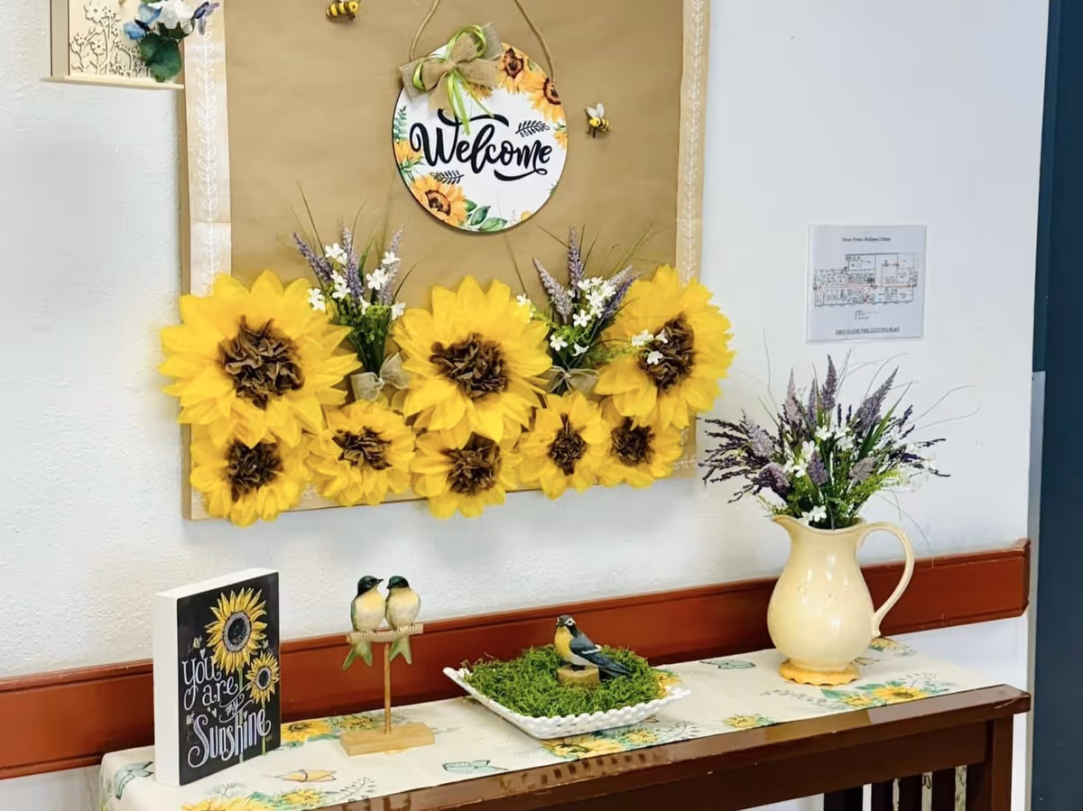 A decorative corner in a wellness center featuring a bulletin board with a 'Welcome' sign surrounded by large artificial sunflowers and small bee decorations. Below the board is a table with a floral tablecloth, a small sign that reads 'You are my Sunshine,' two bird figurines on a stand, a bird figurine on a bed of green moss, and a cream-colored pitcher with purple and white artificial flowers.