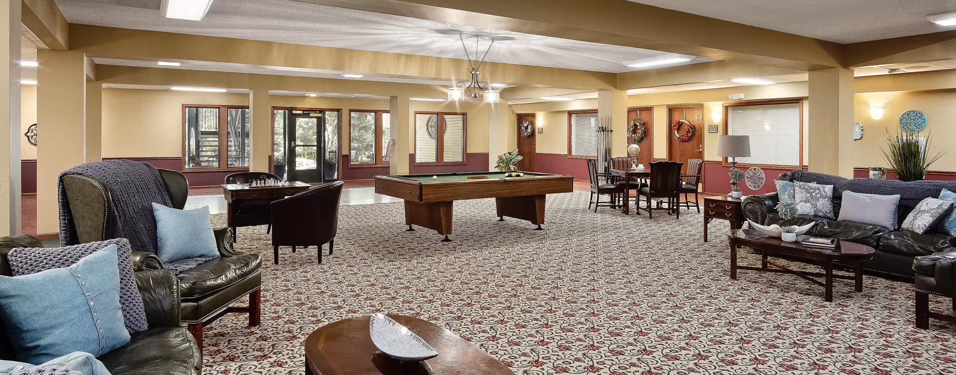 A spacious senior living common area with patterned carpet, leather sofas and armchairs adorned with blue and gray cushions, a pool table in the center, a chess table with chairs, and a dining table with chairs near the back wall. The room has large windows and glass doors letting in natural light, beige walls with maroon wainscoting, and ceiling lights.