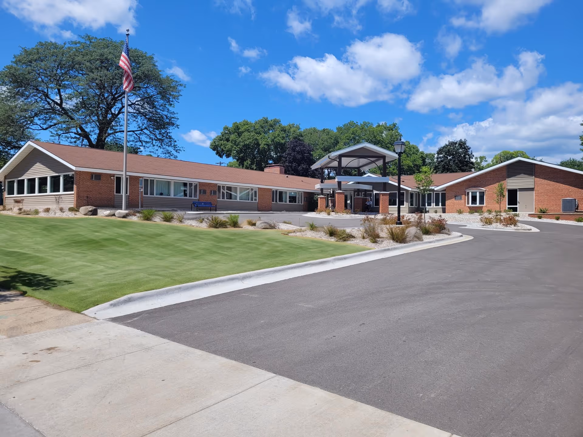 Exterior view of a single-story assisted living facility building with a brick and siding facade, a covered entrance, an American flag on a flagpole, a well-maintained lawn, and a paved driveway under a partly cloudy blue sky.